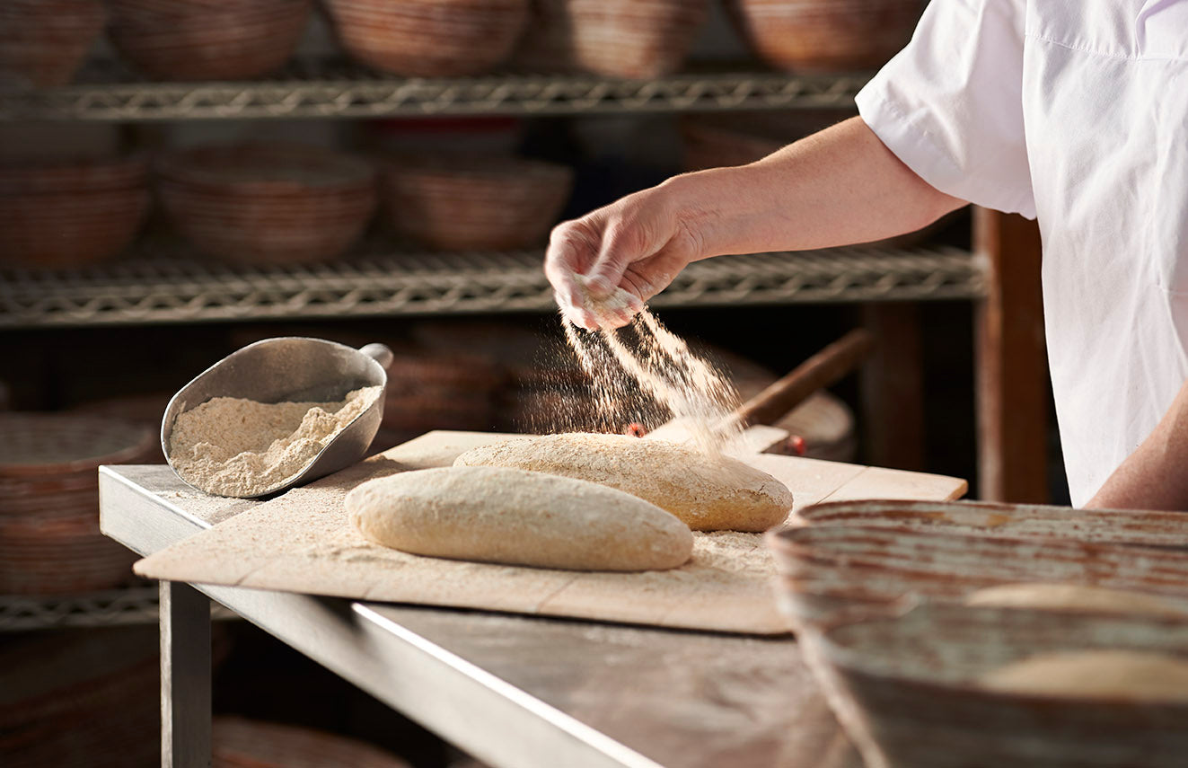 A baker is sprinkling flour on to unbaked loaves of bread. There are wicker bread baskets in the background.