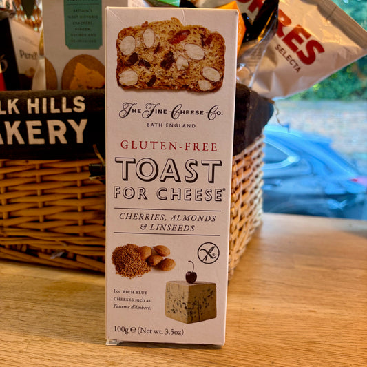 A box of the fine cheese co gluten free toast for cheese on a wooden table in front of a wicker hamper.