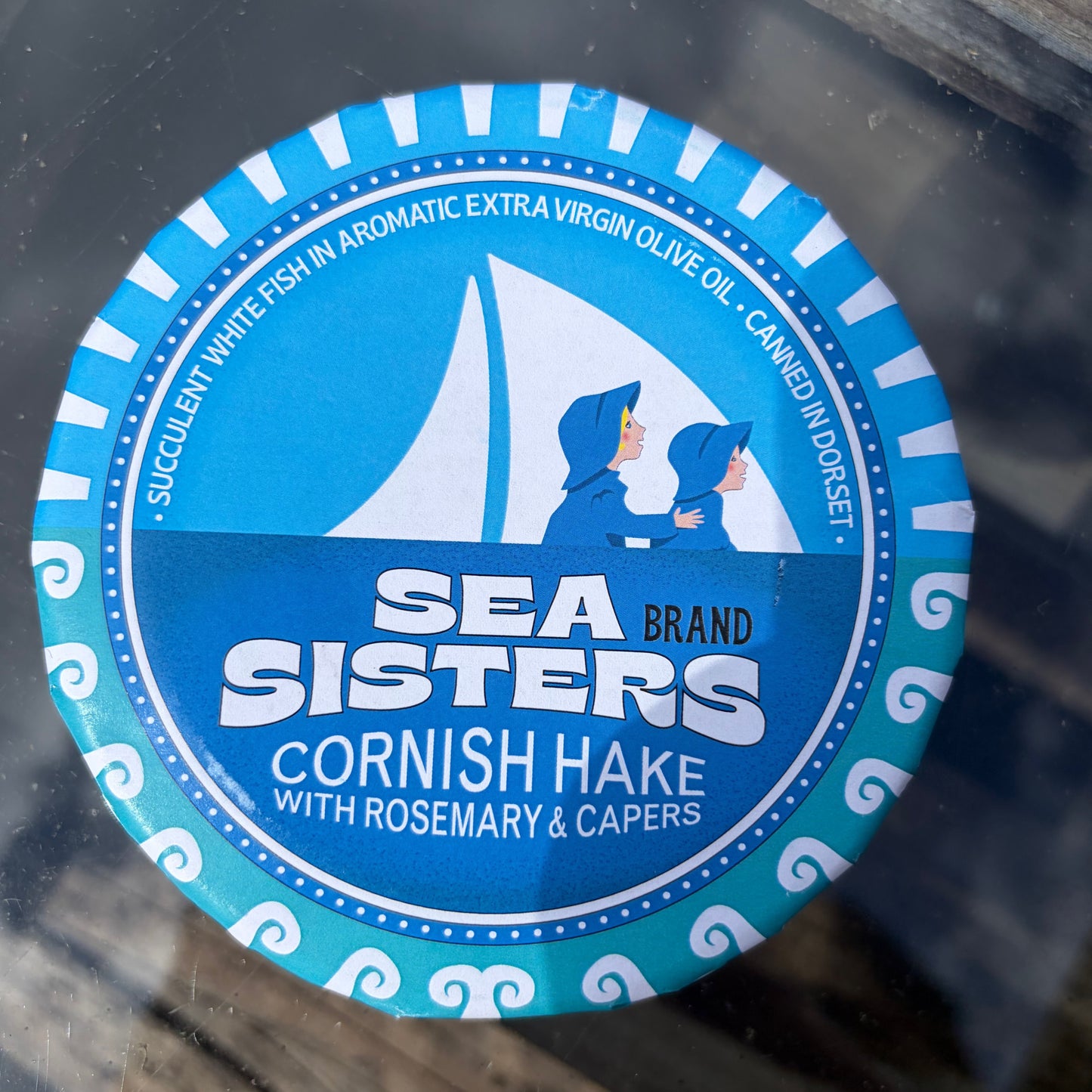 Sea Sisters cornish hake with rosemary and capers tin in blue packaging on glass surface