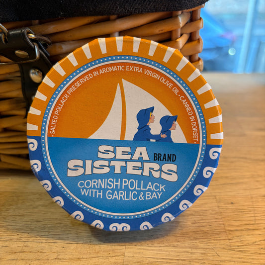 A round metal tin of Sea Sisters Cornish Pollack with Garlic & Bay, resting on a wicker basket next to a dark fabric surface against a wooden background.