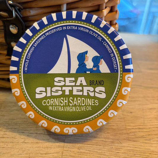 A round metal tin of Sea Sisters Brand Cornish Sardines, featuring a blue, green, and orange label, resting on a wooden surface next to a woven basket.