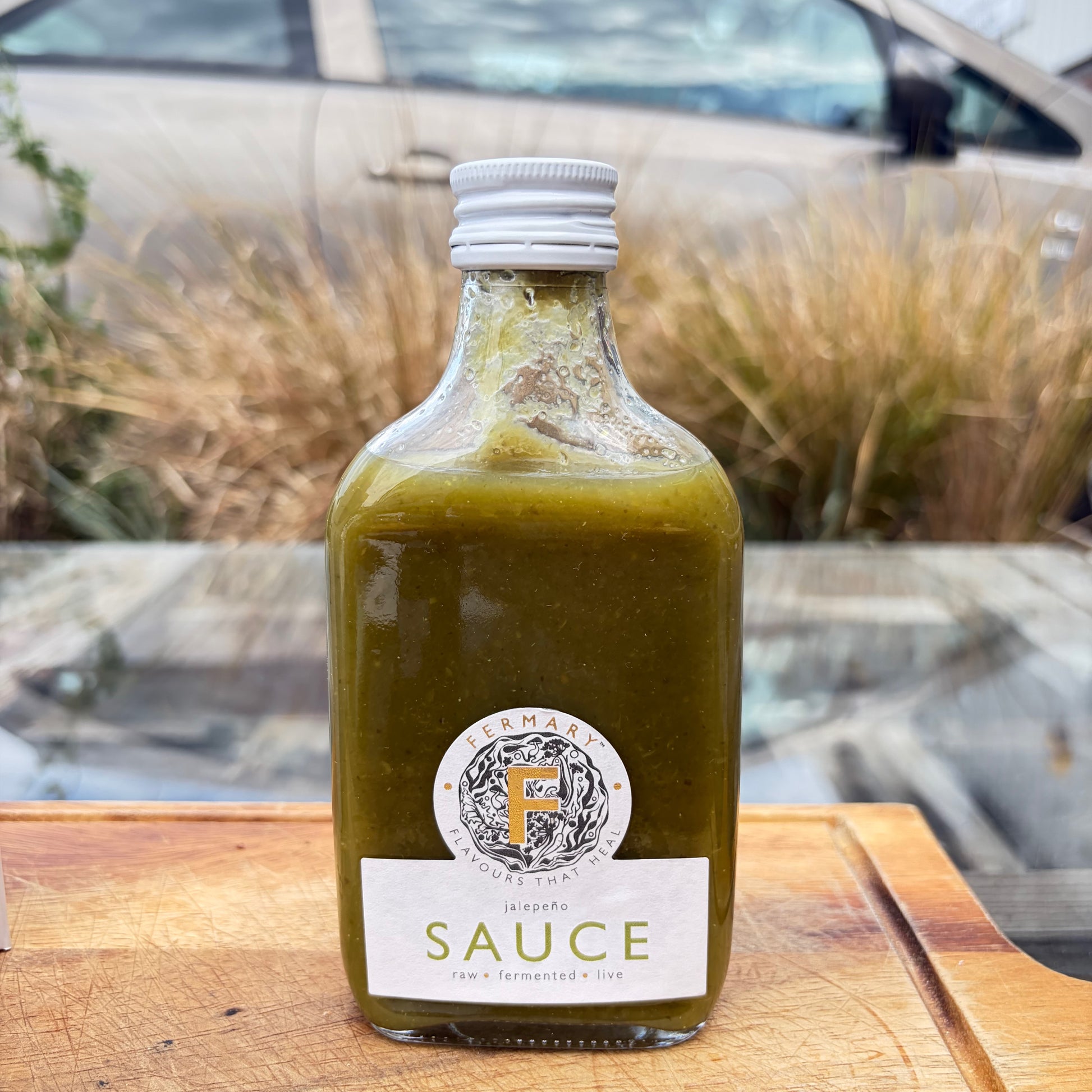 An image of Fermary brand jalapeno sauce placed on a wooden board in front. of grasses.
