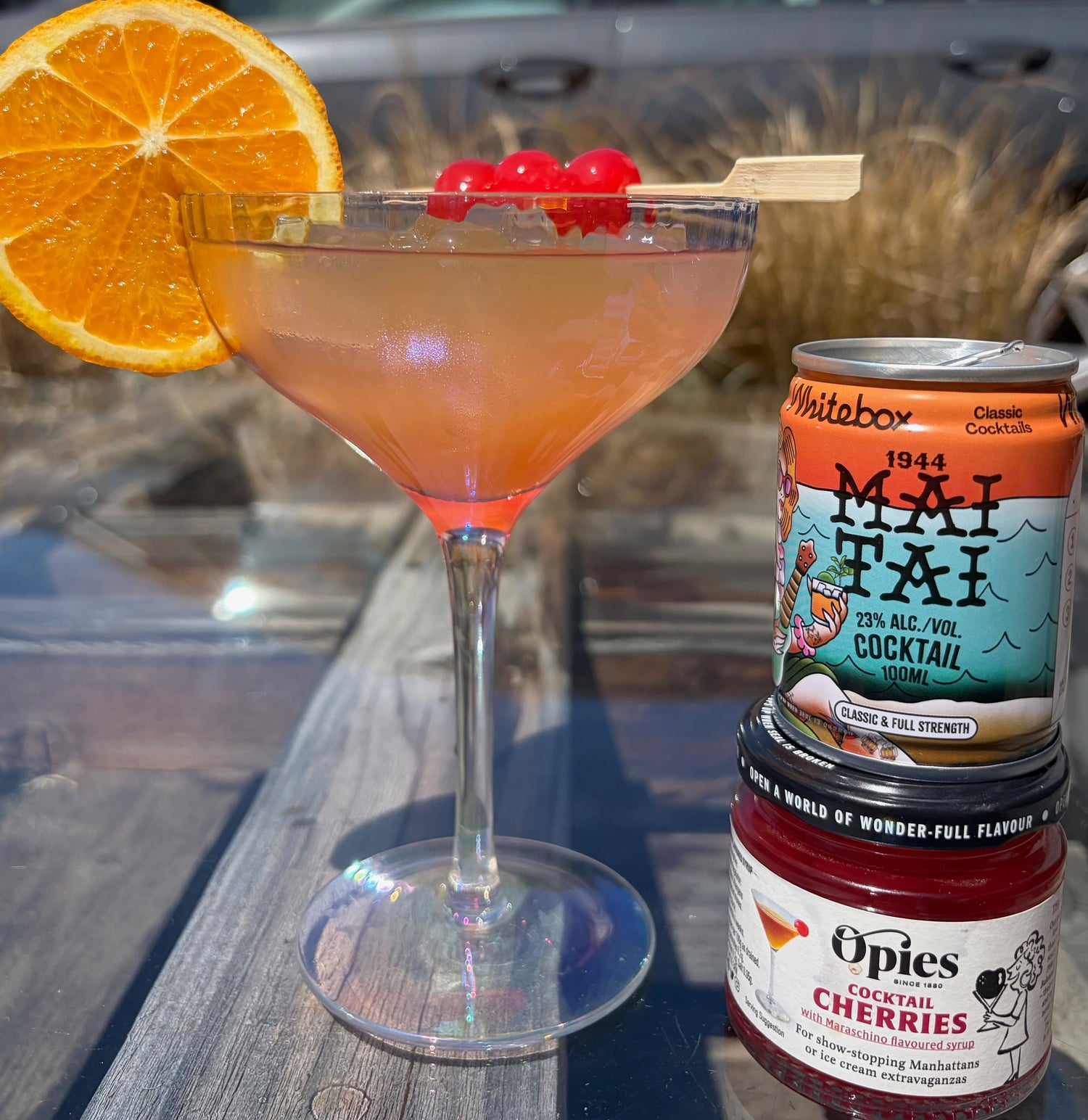 A Mai Tai cocktail in a coupe glass with an orange slice garnish and cherries on a wooden table, next to a can of Whitebox Mai Tai and a jar of Opies Cocktail Cherries.