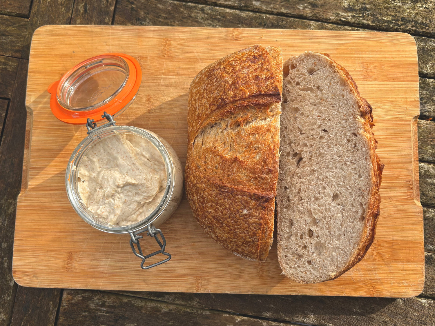 A loaf of rustic sourdough bread, sliced open to show the crumb, arranged on a wooden cutting board next to a small glass jar filled with sourdough starter and an orange-lidded cap, set on a dark wooden table.