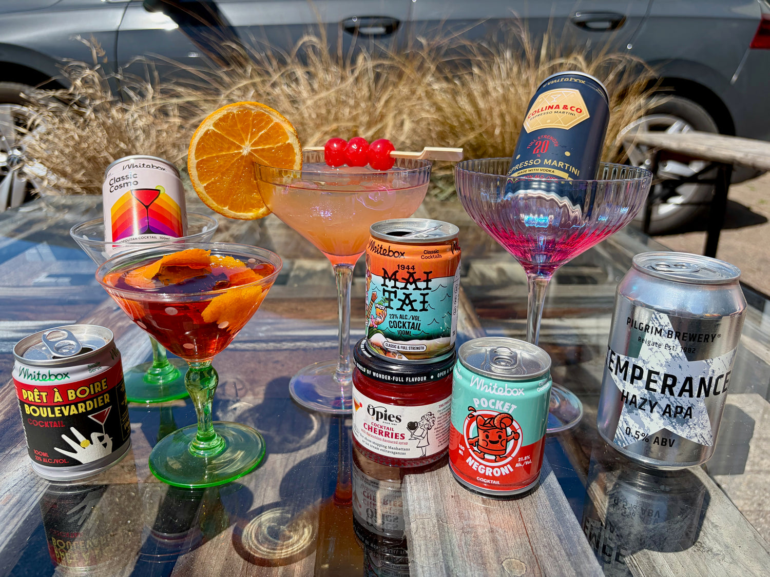 A collection of white box cocktails in glasses and some in cans. There is an orange segment garnish and cocktail cherries on a stick for the Mai Tai. There is a boulevardier in a green and polka dotted glass with an orange zet garnish. There are other cans in glasses not poured out. 