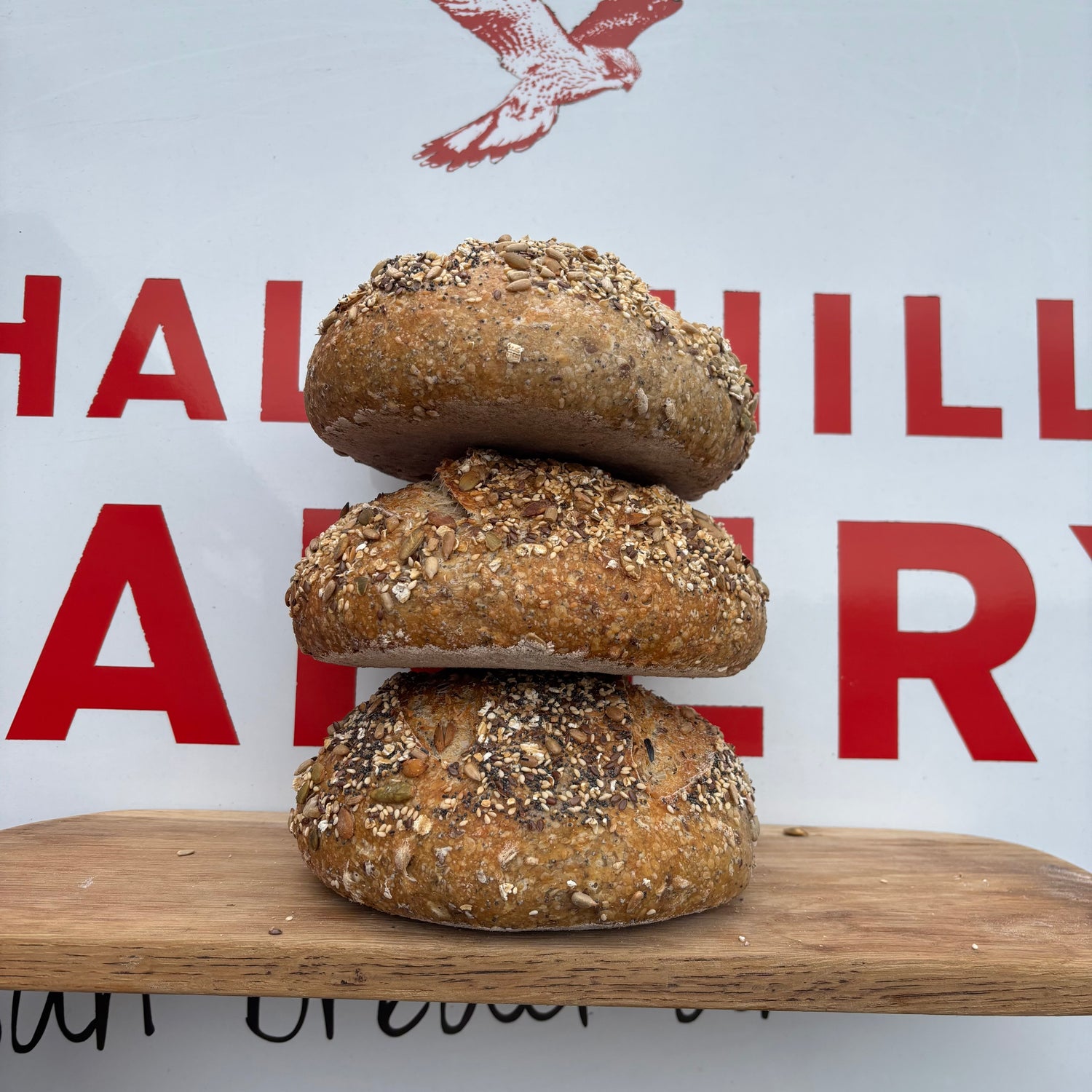 Three stacked artisanal bagels topped with seeds and oats, displayed on a wooden board in front of a white sign with red lettering that reads 'chalk hills Bakery'.